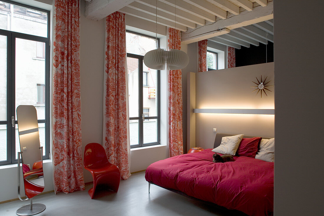Double bed with red cover, red designer chair and cheval mirror in loft apartment