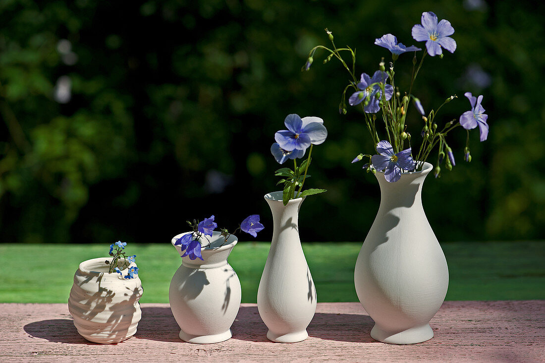Weiße Vasen aufgereiht mit Blüten vom blauen Lein, Hornveilchen, Glockenblume und Vergißmeinnicht