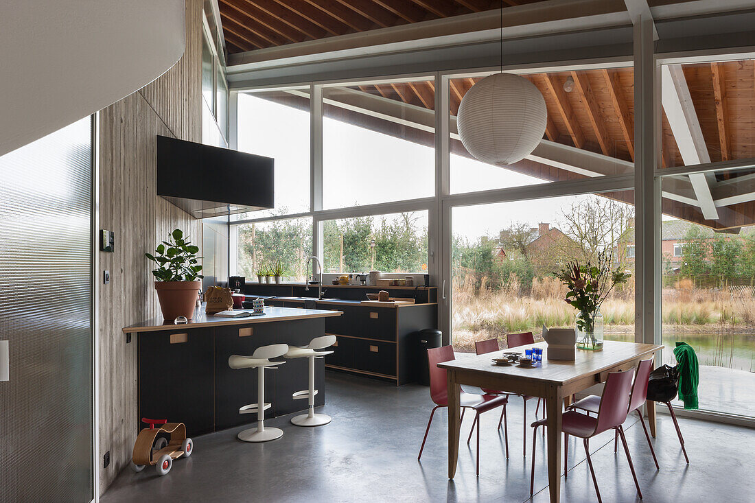 Open-plan kitchen and dining area with large window front, wooden ceiling and concrete wall