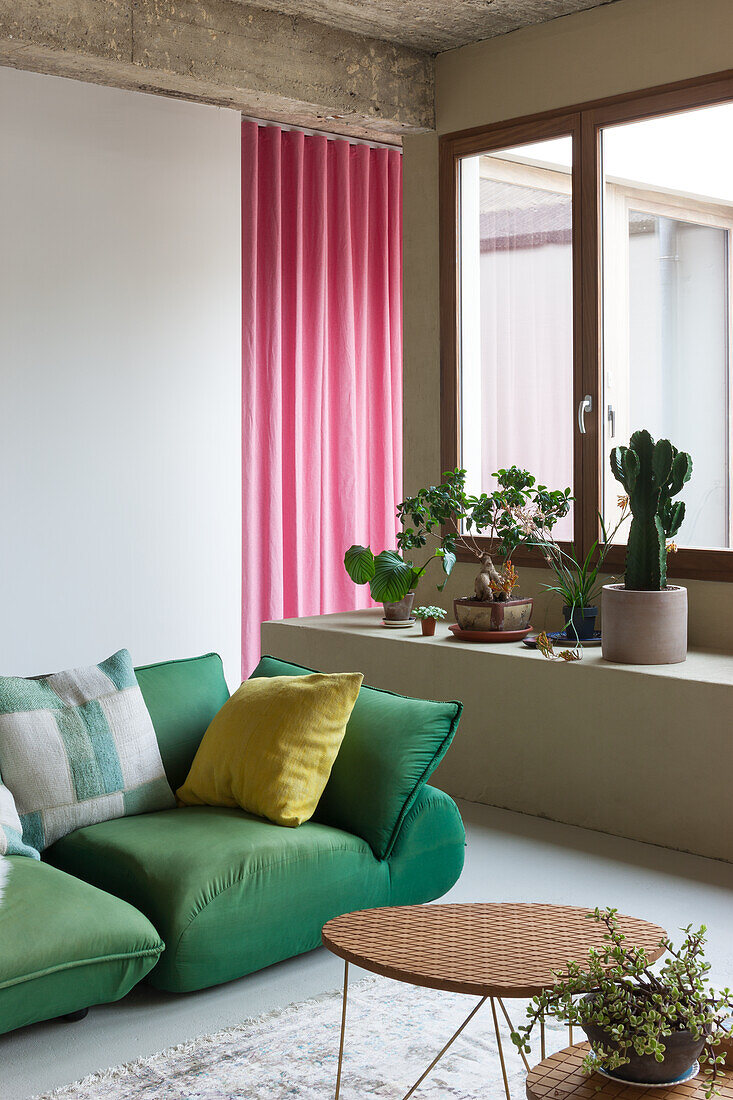 Green sofa with yellow cushions in front of pink curtains, next to window with plants