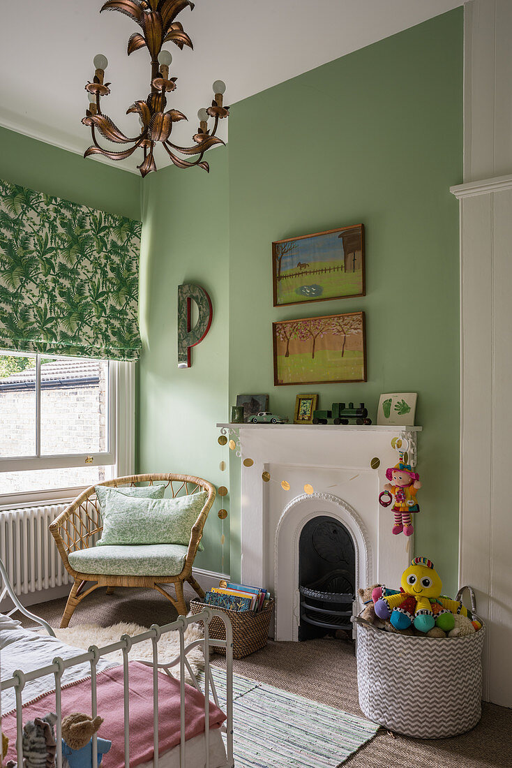 A bamboo chair and a toy basket by a fireplace in a children's room with a green wall