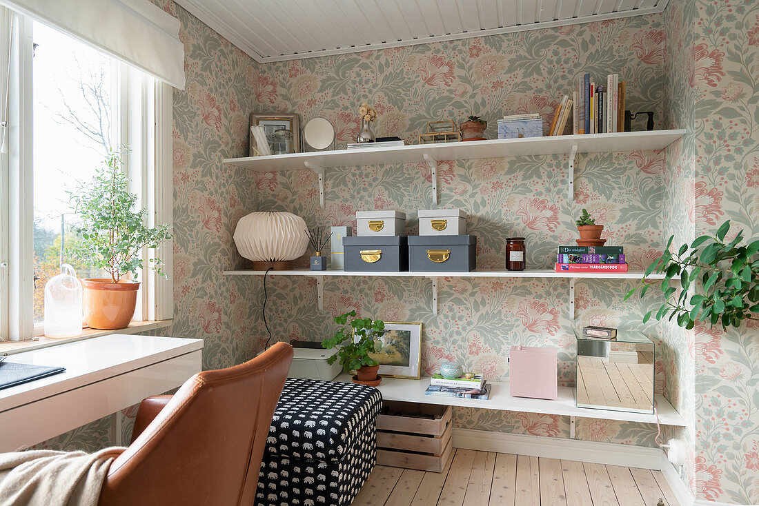 Shelves, an upholstered stool and a desk with a leather chair in a room with floral wallpaper