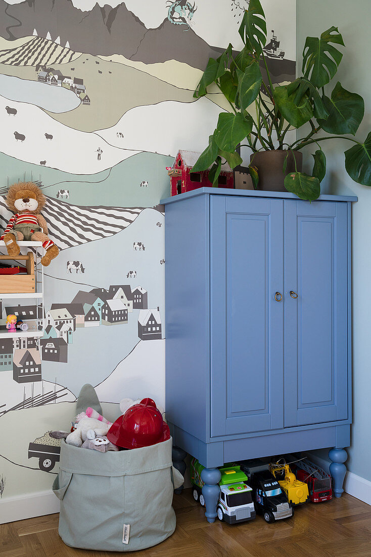 A houseplant on a blue cupboard in a children's room in front of a wall papered with wallpaper with a landscape picture