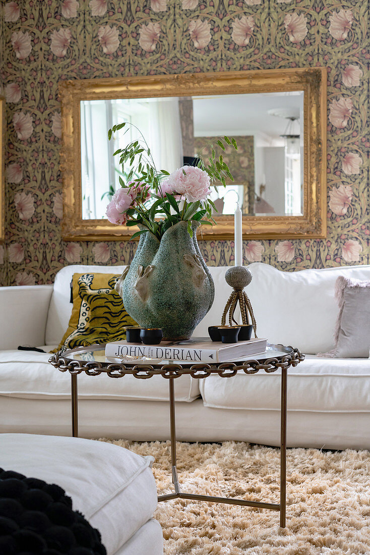 A vase of flowers on a coffee table, a white sofa and a gold-framed mirror on a wall with floral wallpaper