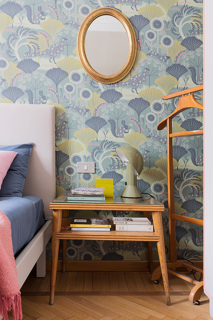 A valet stand and a bedside table next to a bed in a bedroom with wallpaper on the wall