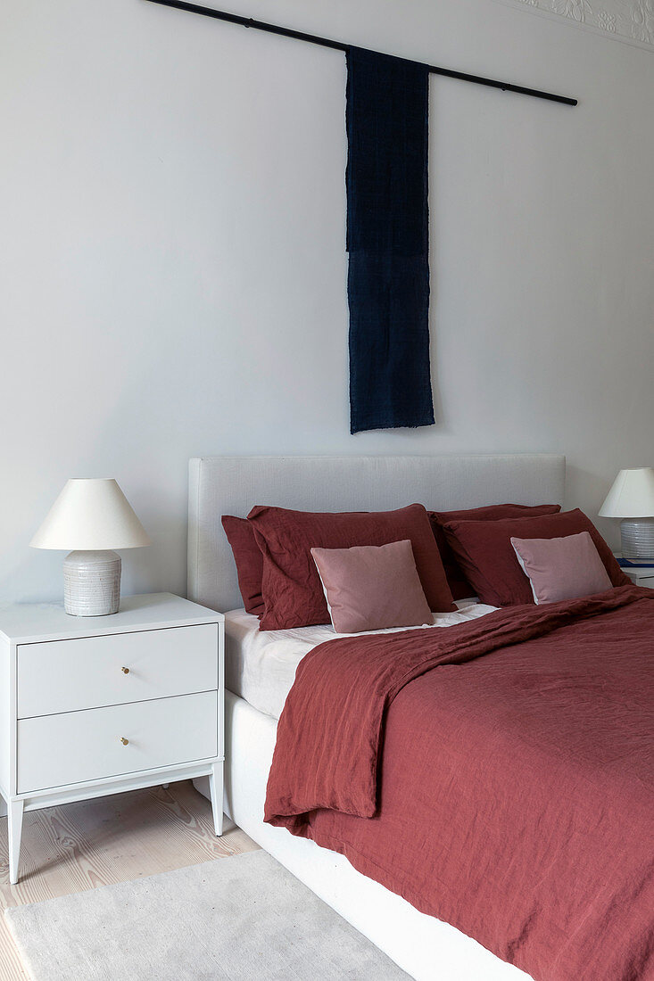 White bed with red bed linen and wall hanging in bedroom
