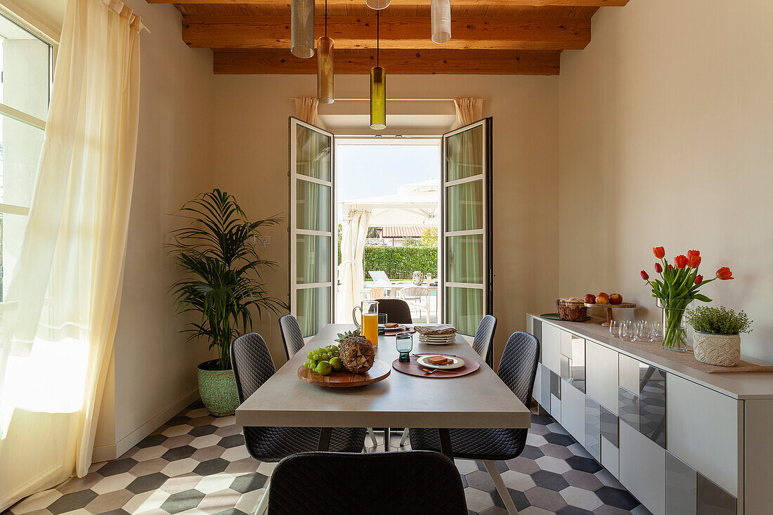 A table with chairs in front of a sideboard in a dining room in front of open patio doors