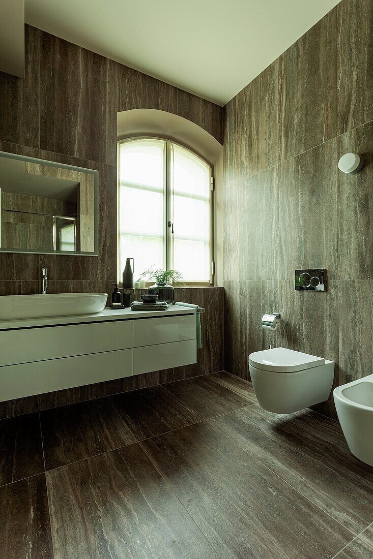A spacious washstand, a toilet and a bidet in a bathroom with dark tiles
