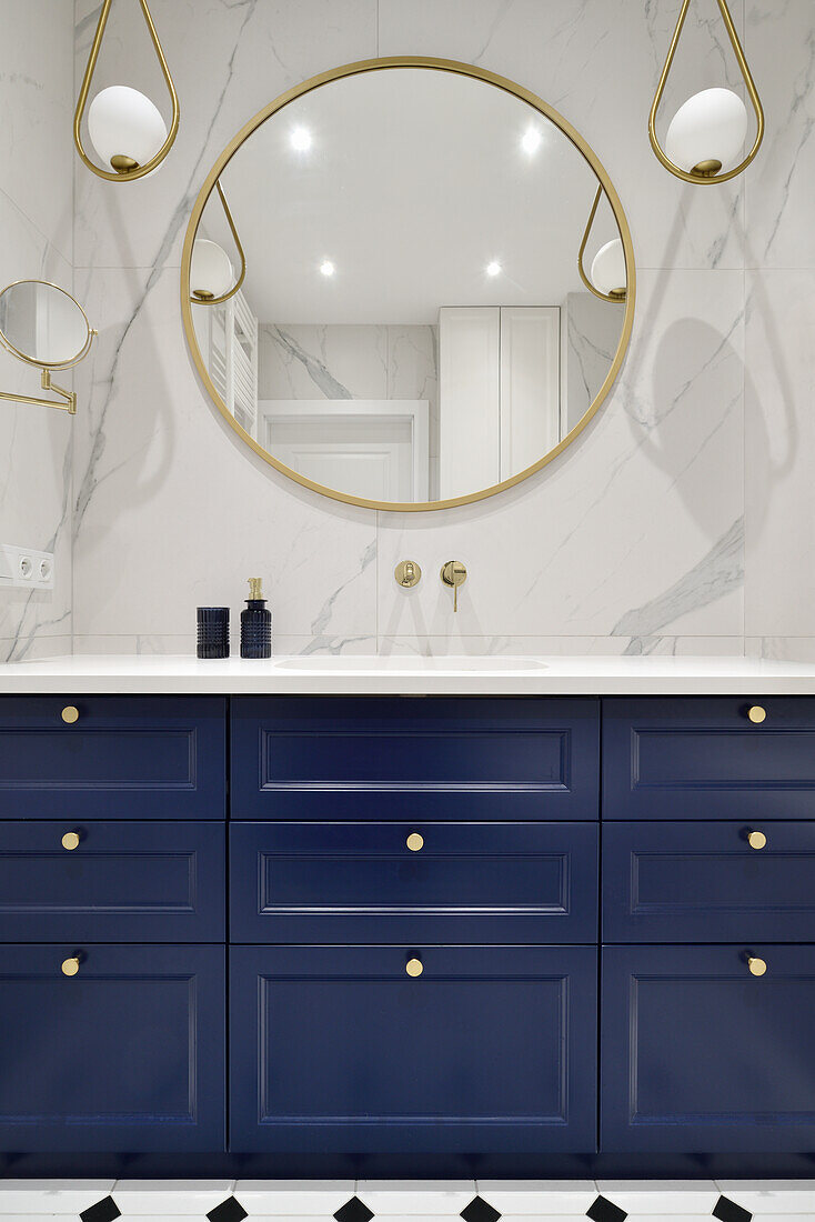 Blue vanity unit, marble tiles above, round mirror and pendant lights in the bathroom