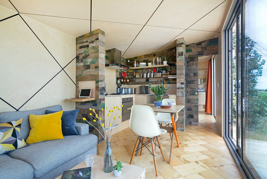 Heller, offener Wohnraum mit grauem Polstersofa, kleinem Essbereich und Küche mit Wänden aus Holzkisten, rautenförmige Decke mit LED-Leisten