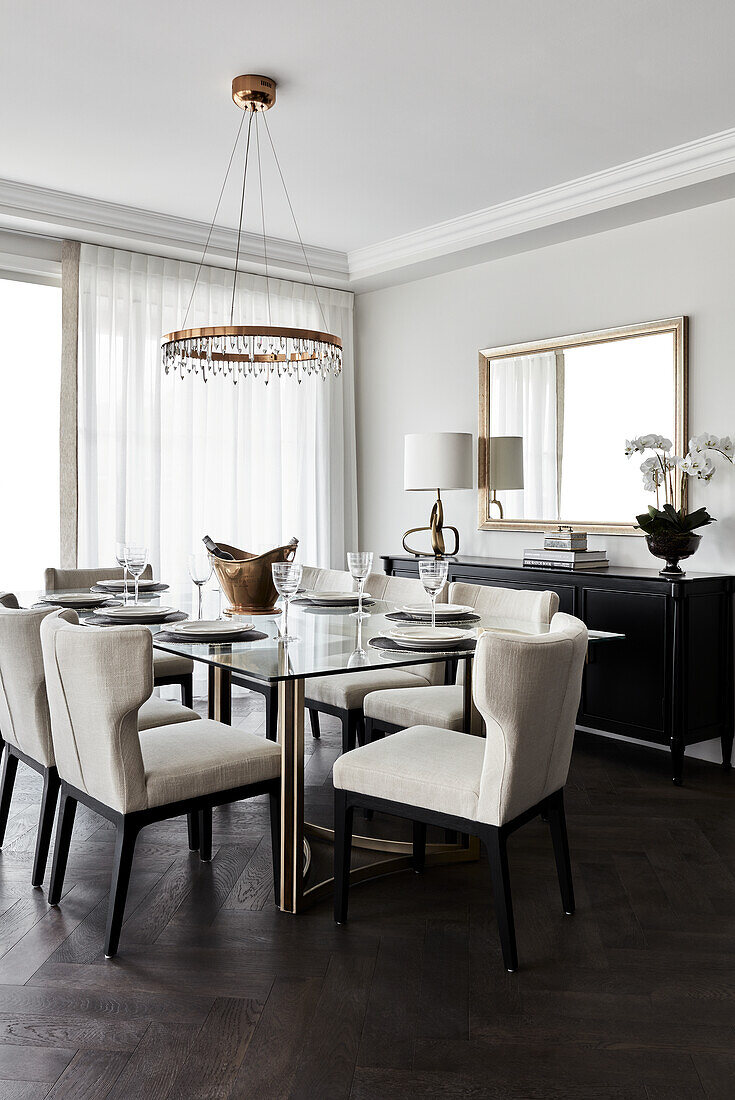 Dining room with glass and brass table, cream upholstered chairs, and chandelier