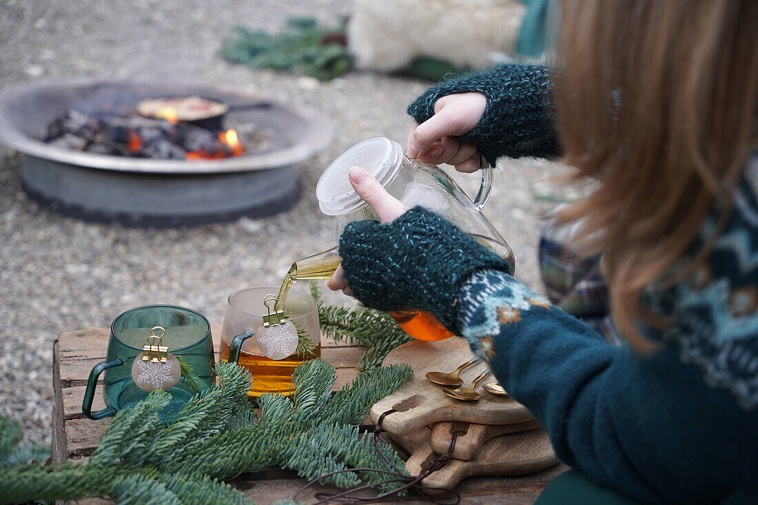 Woman pouring tea in cups with motif made of sustainable wrapping paper with various snowflake motifs in front of fire pit