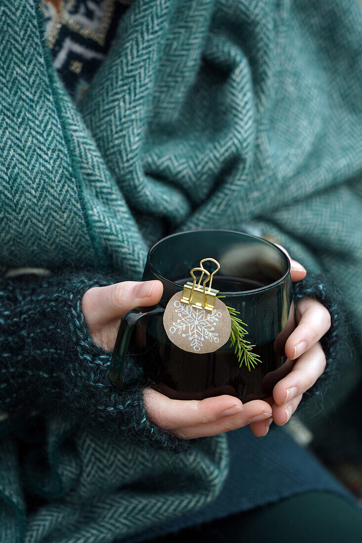 Frau hält Teetasse in den Händen mit Anhänger aus nachhaltigem Geschenkpapier und Schneeflockenmotiven, Wärmen an Teetasse im Freien im Winter