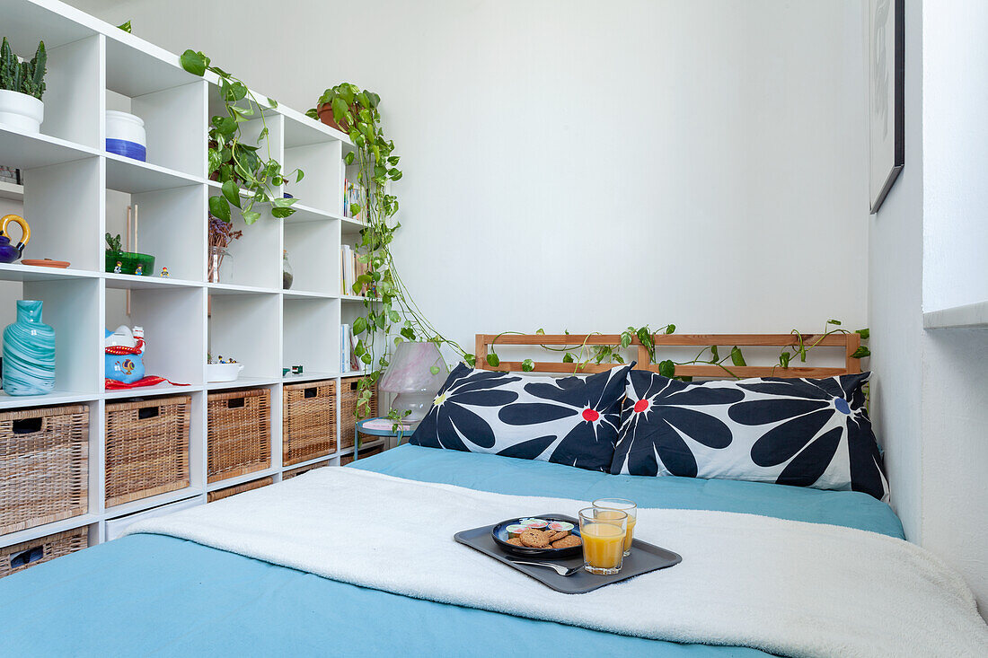 White shelf with baskets and plants next to bed with tray
