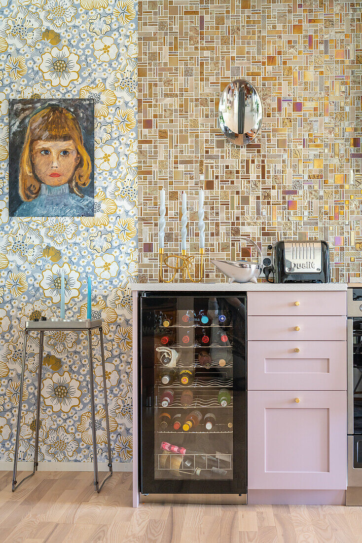 Wallpaper with floral pattern, mosaic tiles, wine fridge and pink-coloured cupboard with drawers