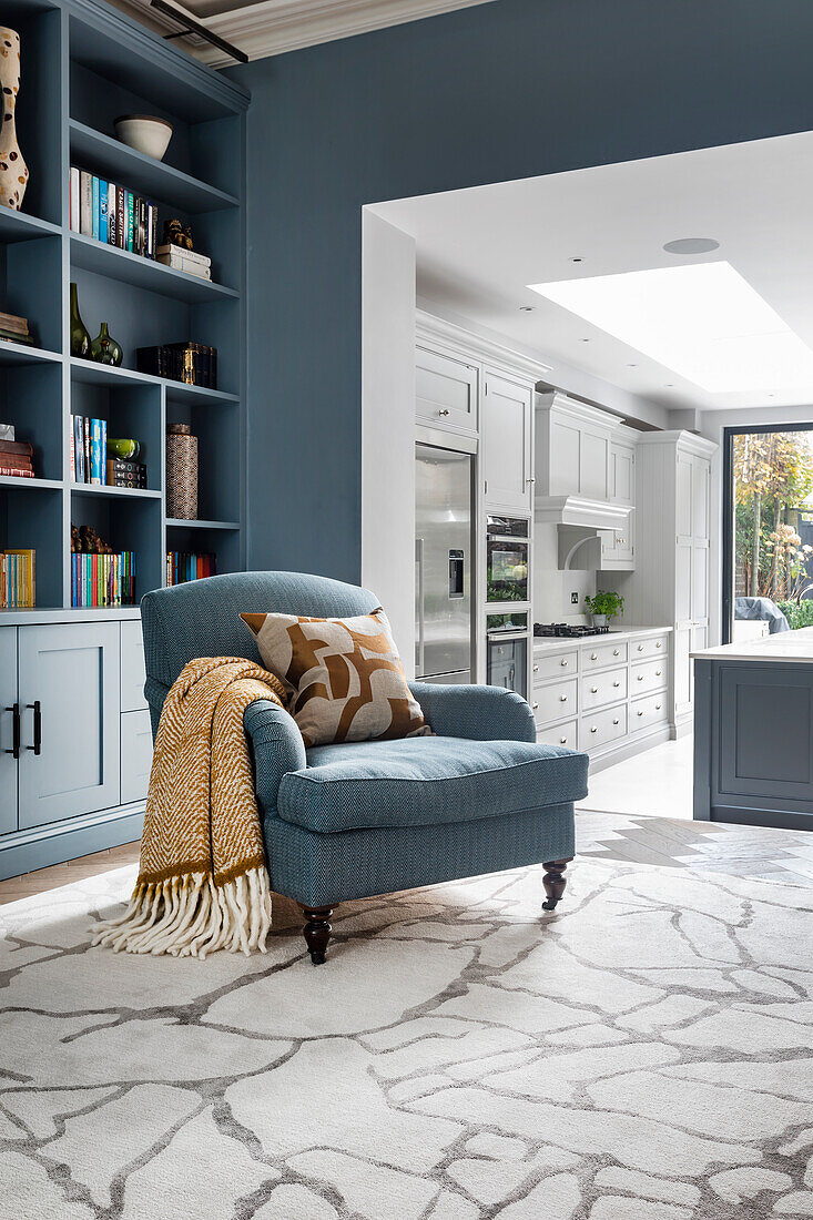 Built-in blue shelving wall, with matching armchair in a living room with light-colored kitchen in the background