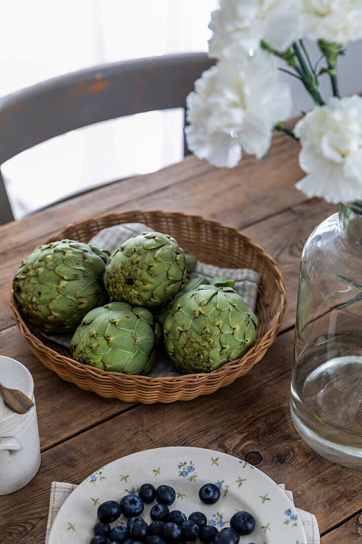 Artichokes in a wicker basket on a wooden table with white flowers and blueberries