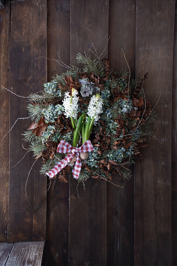 Winter wreath made from natural materials such as larch branches, pine branches, dried hydrangeas, copper beech leaves and fern branches, white hyacinths, hanging on a wooden wall