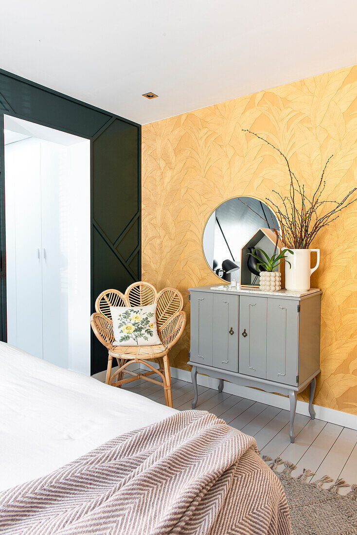 Schlafzimmer mit goldgelber Pflanzen-Tapete, Rattanstuhl und grauer Kommode