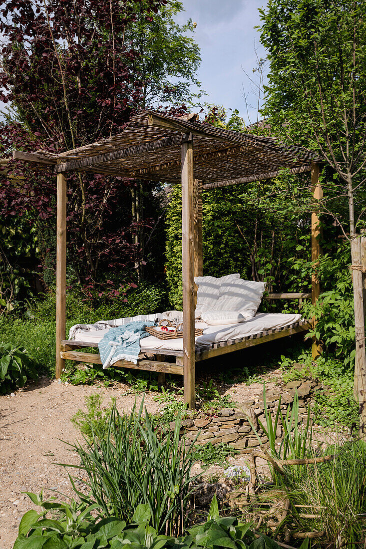 Gartenbett mit Überdachung, Kissen und Decken umgeben von Grünpflanzen