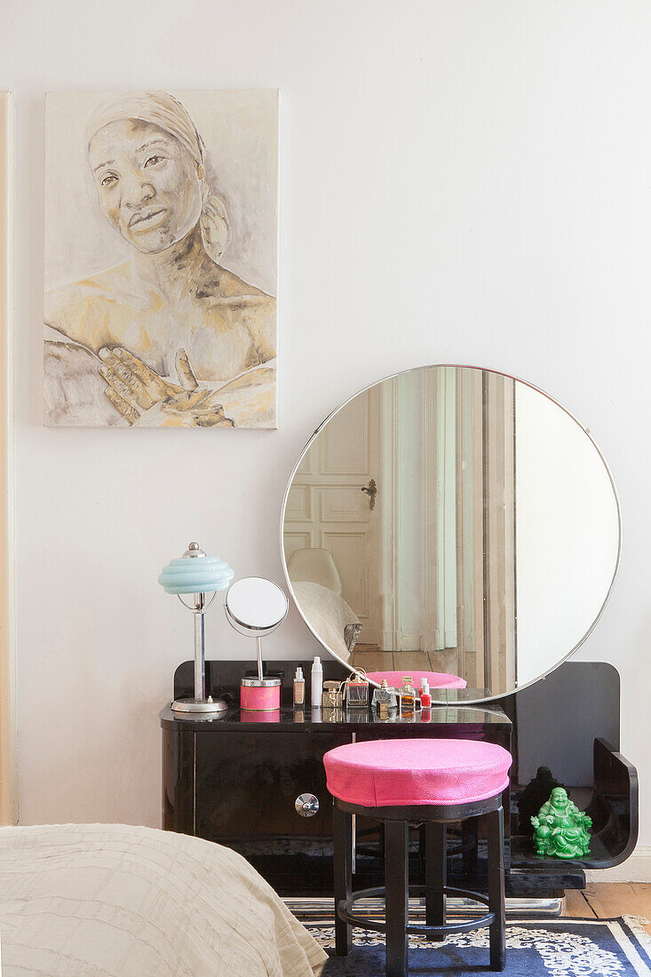 Black dressing table with round mirror and pink stool, portrait above in bedroom