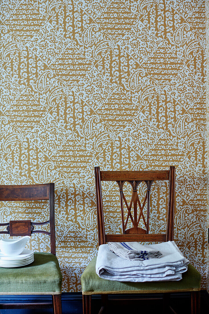 Folded linen and tableware on antique chairs with patterned wallpaper in North Yorkshire farmhouse, UK