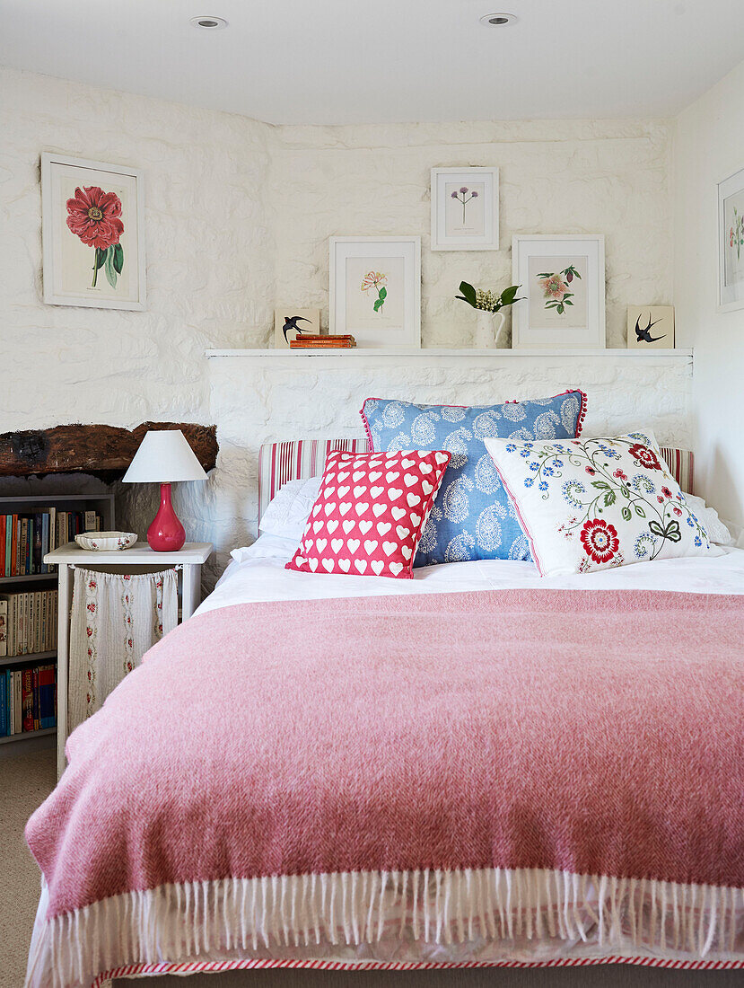 Gemusterte Kissen und gerahmte botanische Drucke mit rosa Decke in einem weiß getünchten Cottage-Schlafzimmer, UK