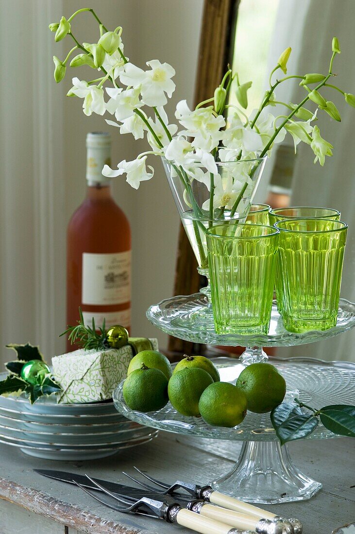 Stillleben mit Glaskuchenständer, Gabeln, Limetten, Blumen und Sellerie, Weinflasche im Hintergrund