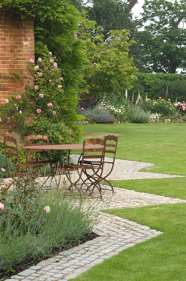 Table with chairs in front of house wall, overgrown with roses and with garden in the background