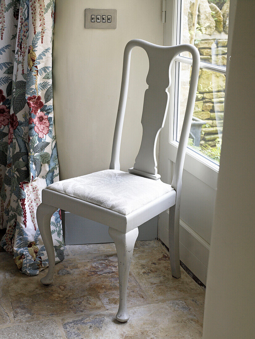 Weiß lackierter Stuhl am Fenster mit geblümtem Vorhangstoff in einem Bauernhaus in Gloucestershire, England, UK