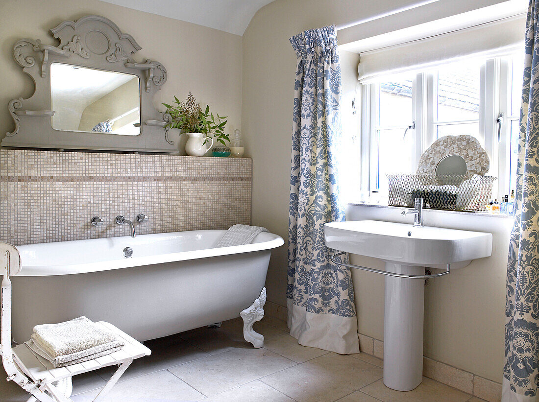 Freestanding rolltop bath with floral curtains in bathroom of Gloucestershire farmhouse, England, UK