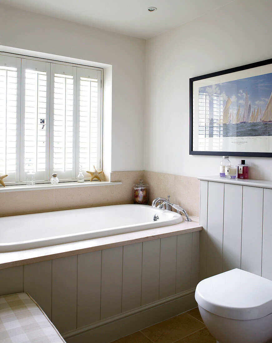 Badewanne unter einem Fenster mit Fensterläden in einem Badezimmer mit nautischem Kunstwerk in einem Bauernhaus in Hampshire, England, UK