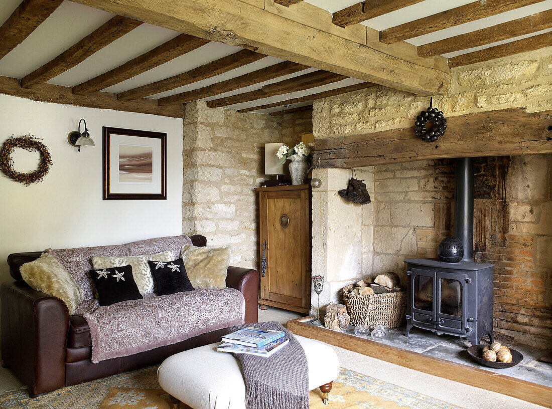 Wolldecken und braunes Ledersofa mit Holzofen im Wohnzimmer eines Bauernhauses in Gloucestershire (England)