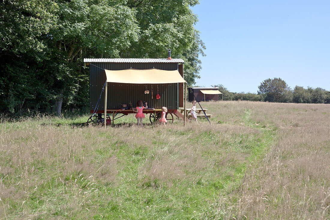 Drei Kinder neben einer Hirtenhütte in einem Campingfeld mit langem Gras