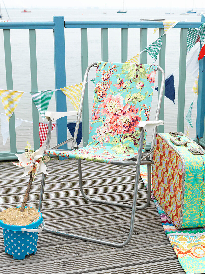 Floral patterned folding chair with vintage suitcase on coastal decking with bunting