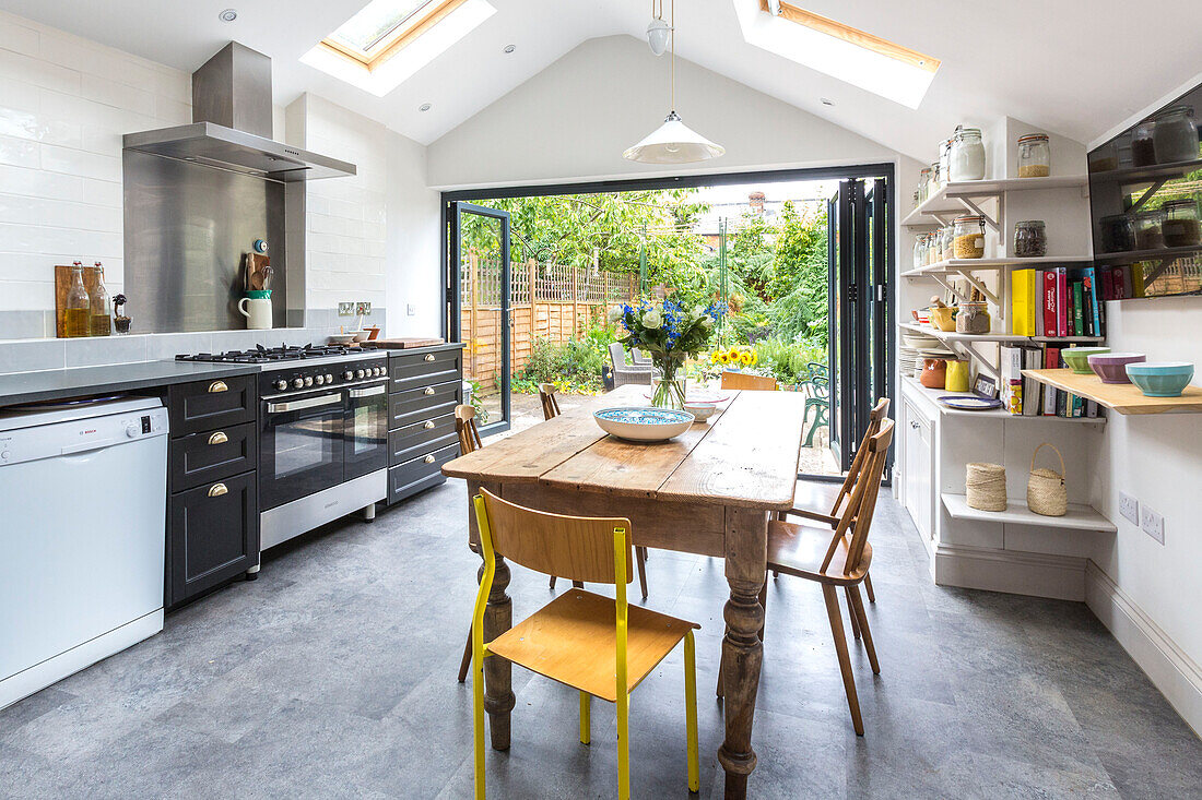 Holztisch und Stühle unter einem Dachfenster mit offenen Türen zur Gartenterrasse in einer Küche in Reading, Berkshire, England UK