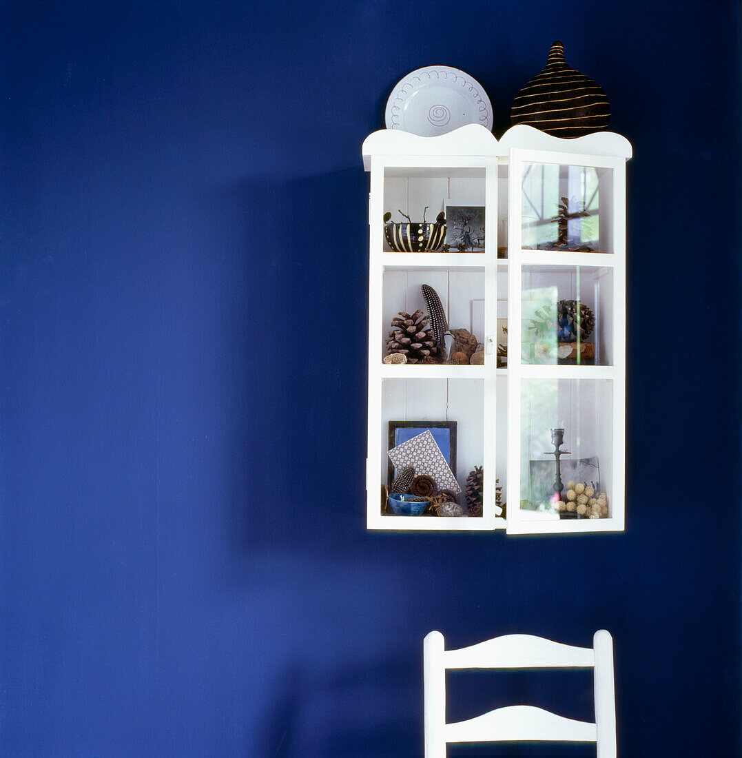Blau gestrichene Wand mit weiß gestrichenem Schrank, gefüllt mit natürlichen Fundstücken, und weißem Stuhl