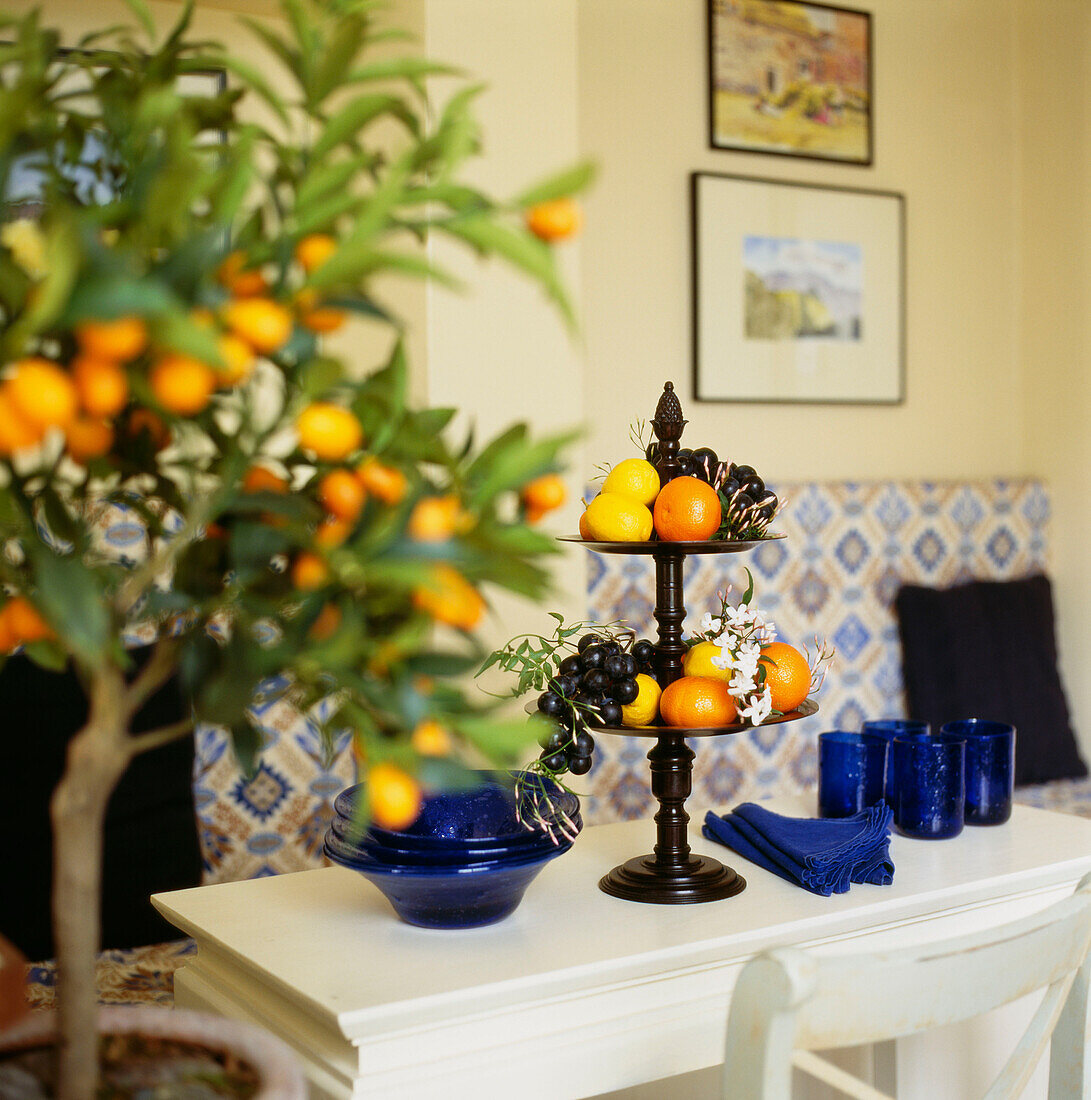 Obst mit Jasminblüten auf einem Holzständer, Orangenbaum im Vordergrund und blaue Gläser auf einem Tisch neben einer gepolsterten Bank