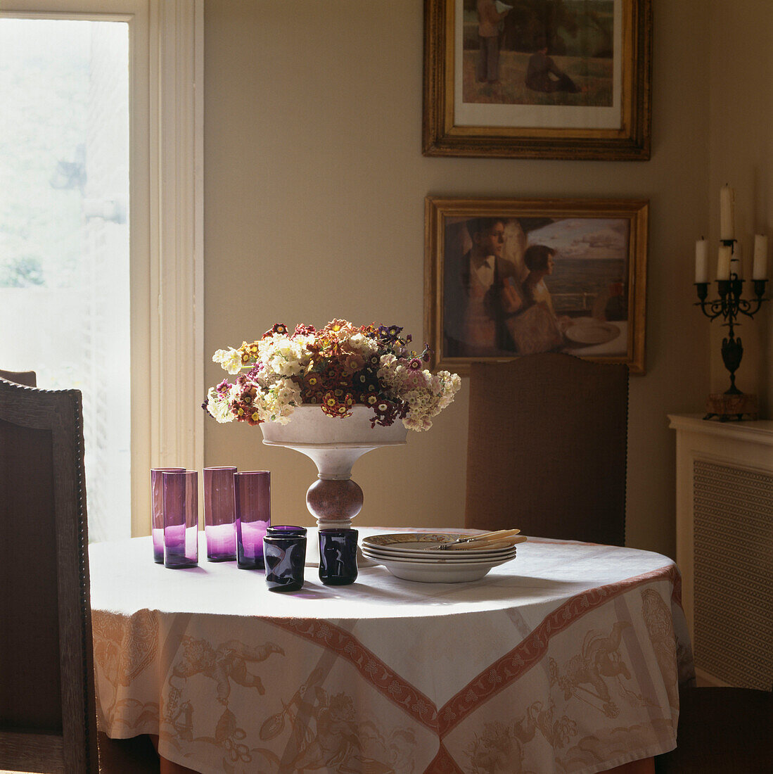 Esstisch mit Tischtuch und lilafarbenen Gläsern und Tafelaufsatz mit Aurikularblumen neben dem Fenster