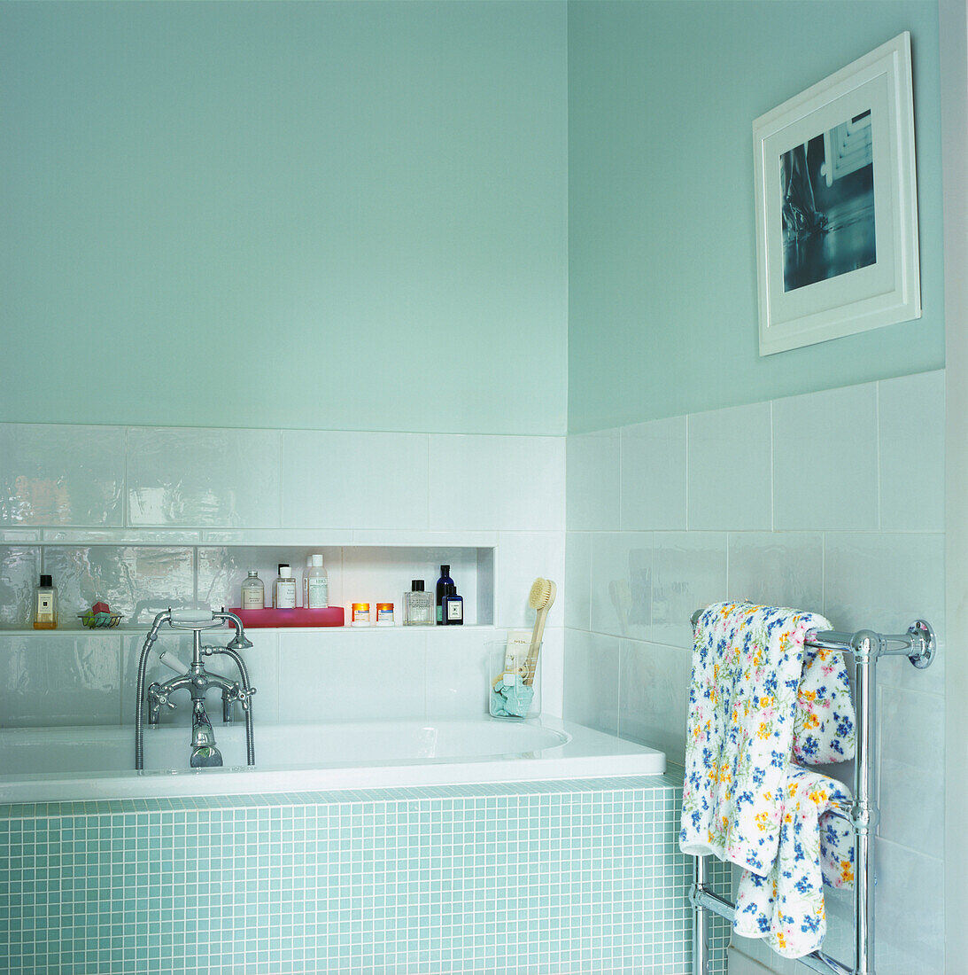 Pale blue bathroom with mosaic tiled bath surround and recessed shelf with toiletries and heated towel rail