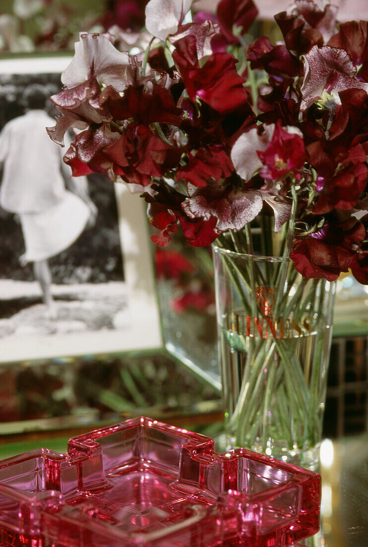 Glas mit roten und violetten Duftwicken auf Tisch mit Aschenbecher und gerahmtem Foto