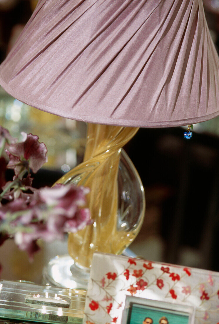 Detail eines Tischlampenständers aus gedrehtem Glas mit einem Lampenschirm aus rosa Seide mit Blumen und einem Foto im Vordergrund