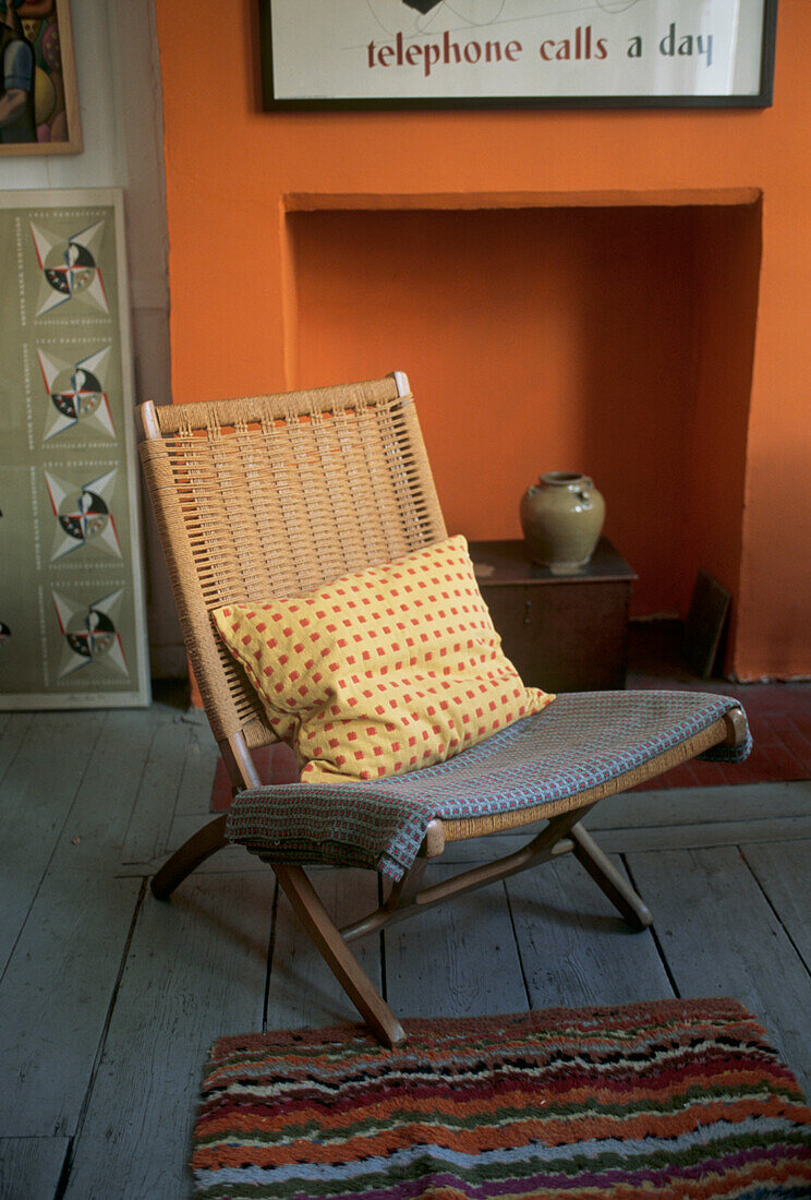 Klappstuhl aus gewebtem Seegras auf grauem Dielenboden vor einer orangefarbenen Wand