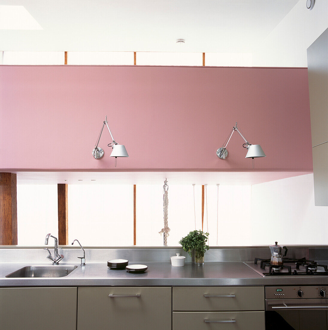 Moderne Küche mit freitragenden Wandleuchten aus Aluminium an einer auffallend rosafarbenen Wand über einer Arbeitsplatte aus Edelstahl auf grauen Einbauschränken