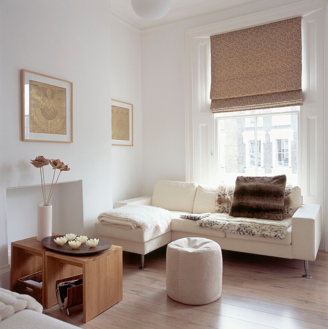 Neutrales Wohnzimmer mit cremefarbenem Ecksofa mit Wolldecken und Kunstfellüberwurf vor einem großen Schiebefenster mit Raffrollos