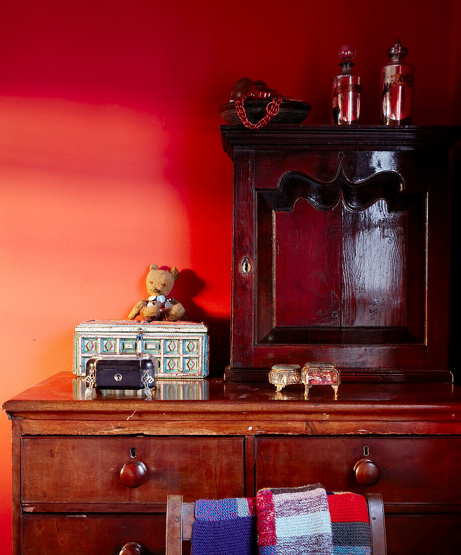 Nahaufnahme von Vintage-Möbeln aus dunklem Holz vor einer tiefrot dekorierten Wand mit Schmuckkästchen und Erinnerungsstücken im Schlafzimmer