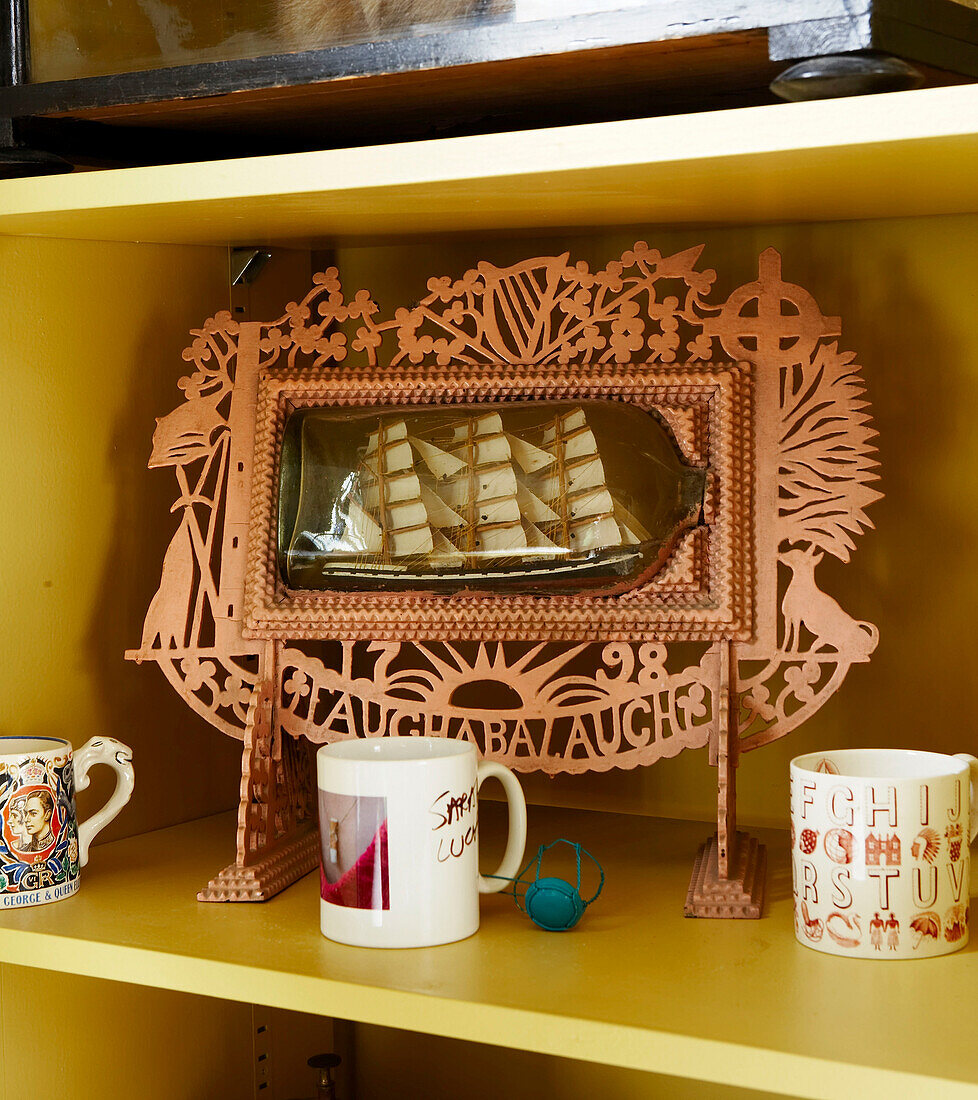 Decorative stand in folk art style with a ship in a bottle on a shelf with some mugs