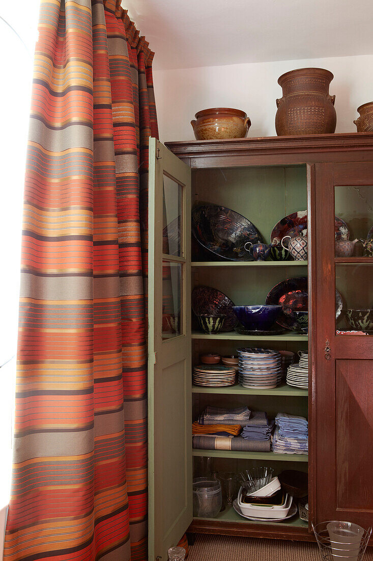 Upcycled vintage storage cupboard full of crockery and tableware next to stiped curtain