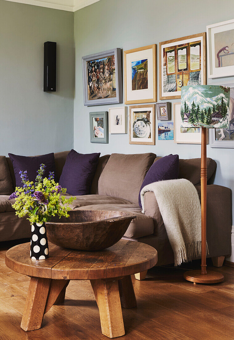 Kunstwerke über einem braunen Sofa mit lila Kissen und einem niedrigen Holztisch im Wohnzimmer eines Familienhauses in Rye, East Sussex, England, UK