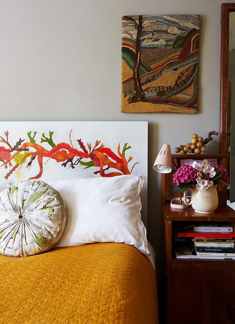 Artwork above hand-painted headboard with cut flowers on side cabinet in Hackney bedroom, East London, UK